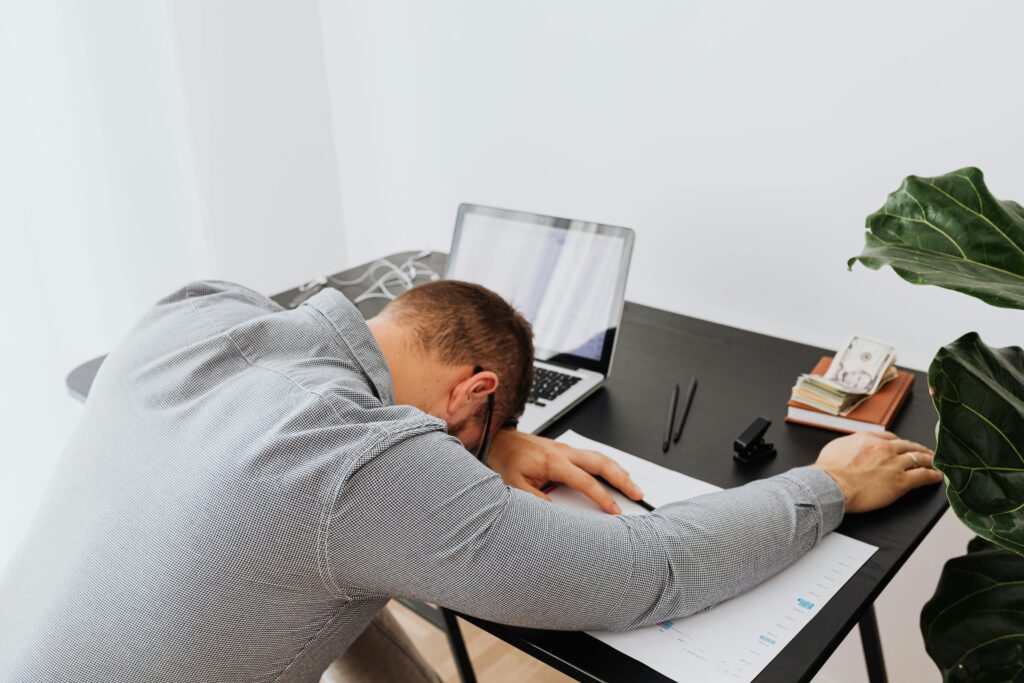 pexels-photo-5717792-5717792 Tired man asleep at a desk with an open laptop and paperwork, indoors.