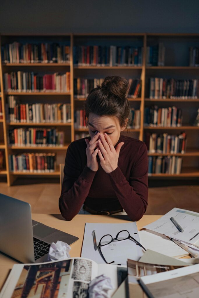 pexels-photo-8085935-8085935 Woman feeling stressed while studying in a library, surrounded by books and laptop.
